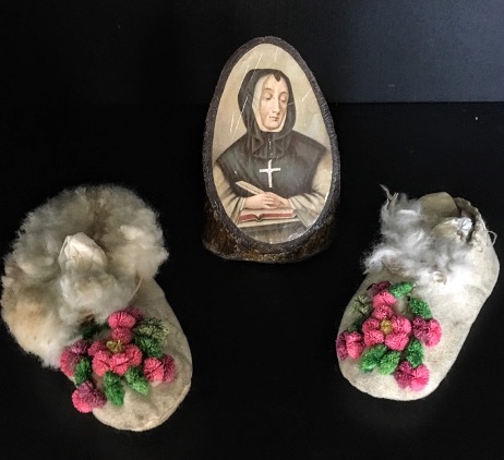 A picture containing two baby-sized mocassins and a picture of a nun superimposed on a cut of a tree branch.

Description automatically generated